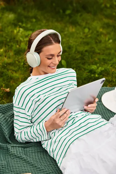 A young beautiful woman relaxes on a picnic blanket, enjoying music and a tablet outdoors. — Stock Photo