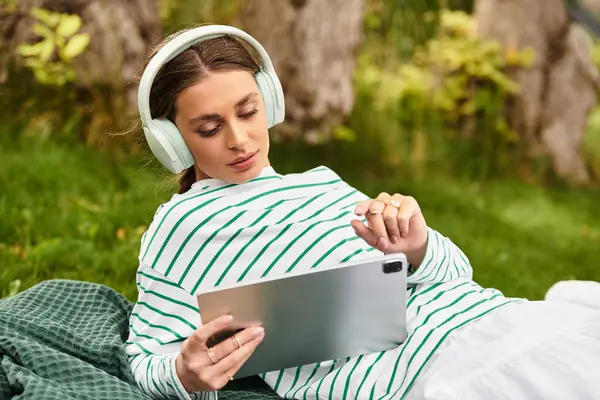 A beautiful young woman in headphones relaxes on a picnic blanket while using a tablet outside. — Stock Photo