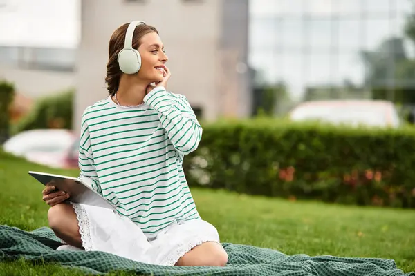 Una joven hermosa mujer se sienta en una manta, disfrutando de su picnic mientras escucha música al aire libre. - foto de stock