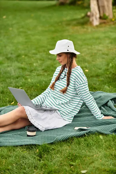 Hermosa mujer con un sombrero se sienta en una manta de picnic, se centró en su computadora portátil al aire libre. - foto de stock