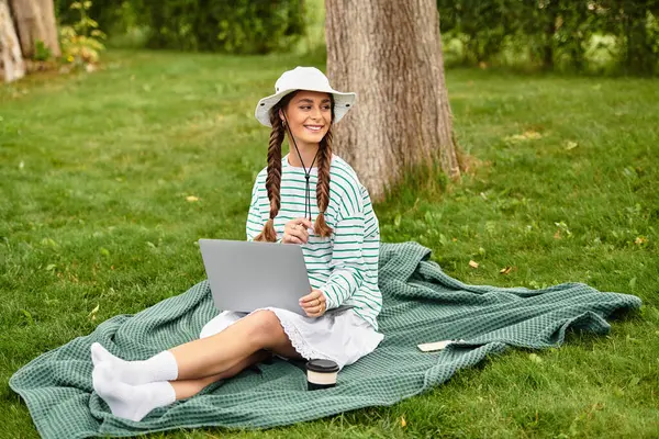 A young beautiful woman sits on a picnic blanket, smiling while using a laptop in a peaceful park. — Stock Photo