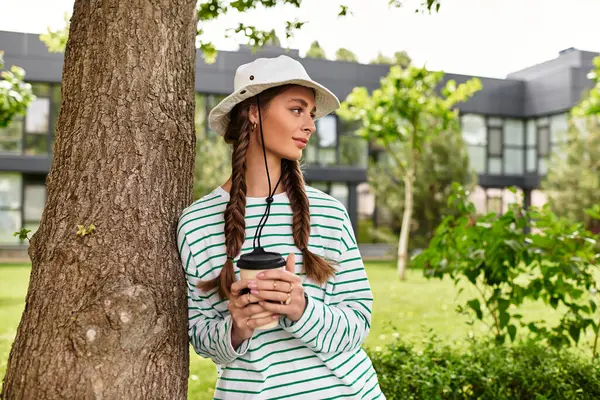 A young beautiful woman savors a warm drink while peacefully resting against a tree in the park. — Stock Photo