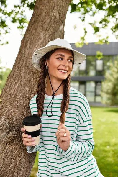Una joven con el pelo trenzado sonríe brillantemente mientras sostiene una taza de café, relajándose en el parque. - foto de stock