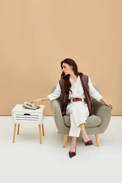 A confident young woman with long brown hair sits elegantly in a cozy armchair, dressed stylishly. — Stock Photo