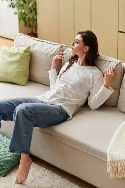 A beautiful young woman with brown hair relaxes on a cozy couch, savoring a quiet moment. — Stock Photo
