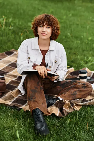 A young woman with curly hair sits on a blanket, writing in her notebook at a vibrant park. — Stock Photo