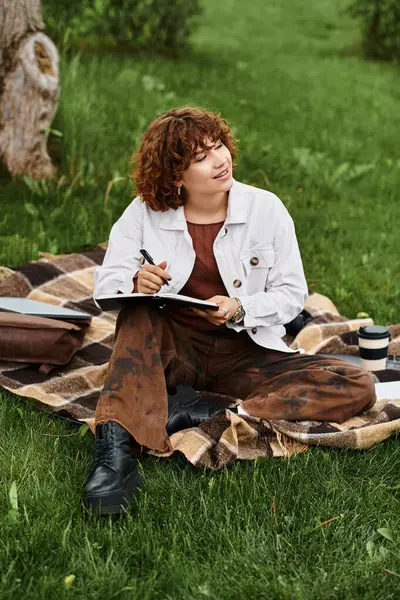 A young woman with curly hair sits on a blanket in a park, sketching and enjoying summer. — Stock Photo