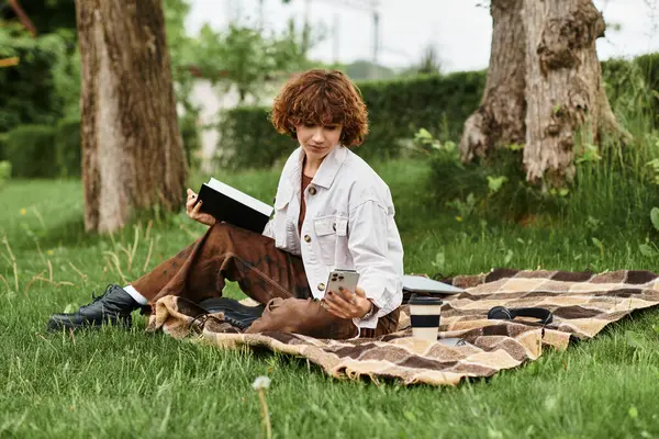 Young woman enjoys a sunny day on a blanket while reading and checking her phone in the park. — Stock Photo