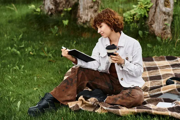 Curly-haired woman sits on a blanket, sipping coffee while writing in her notebook in the park. — Stock Photo
