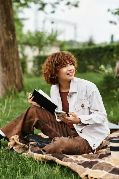 A young woman with curly hair enjoys reading while seated on a blanket in a lush park. — Stock Photo