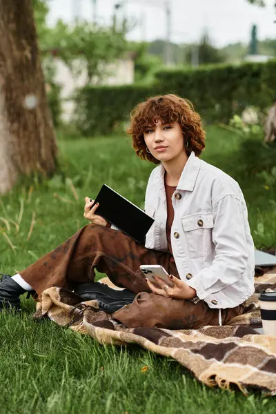 Curly-haired young woman relaxes on a blanket, jotting notes in her notebook amidst summer greenery. — Stock Photo