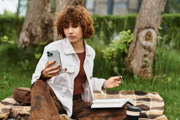 Curly-haired woman lounges on a blanket, absorbed in her phone while surrounded by nature beauty. — Stock Photo