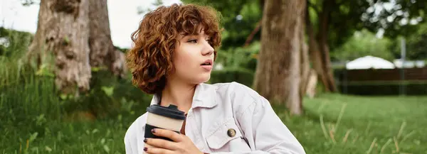A young woman with curly hair relaxes in the park, sipping coffee in the sun, banner — Stock Photo