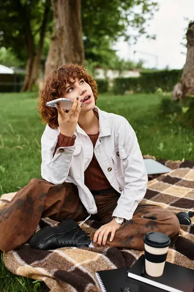 Curly-haired young woman sits on a blanket in a park, conversing casually on her phone. — Stock Photo