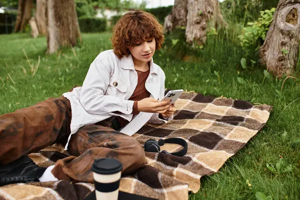 Young woman lounges on a blanket in a green park, engrossed in her smartphone. — Stock Photo