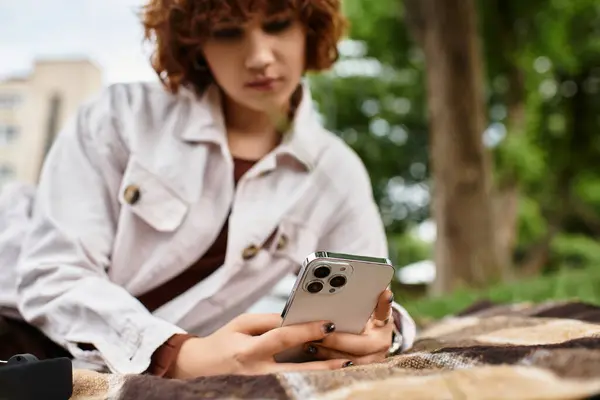 Relaxing in the park, a young woman with curly hair focuses on her phone while seated. — Stock Photo