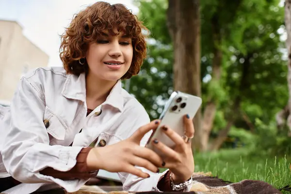 Curly-haired woman relaxes on a blanket while looking at her phone in a sunny park. — Stock Photo
