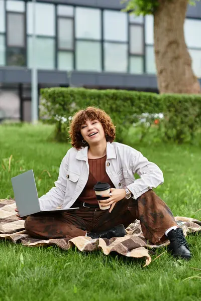 Curly-haired young woman works outdoors on her laptop while enjoying coffee in a vibrant park. — Stock Photo
