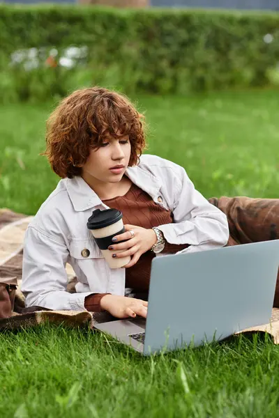 A young woman with curly hair is working on her laptop in a green park during summer. — Stock Photo