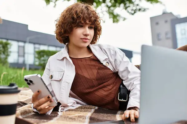 A young woman with curly hair enjoys working remotely on a blanket in a lush park. — Stock Photo
