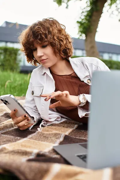 Curly-haired woman relaxes on a blanket while working remotely in a sunny park. — Stock Photo