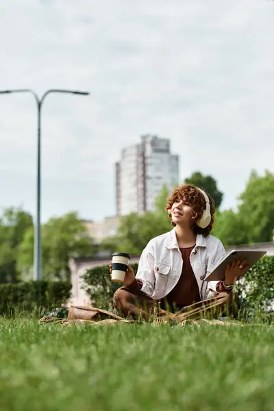 Curly-haired woman sits cross-legged on a blanket, sipping coffee and using a tablet outdoors. — Stock Photo