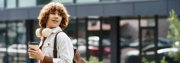 A young woman with curly hair stands outdoors holding coffee and smiling near a contemporary office, banner — Stock Photo