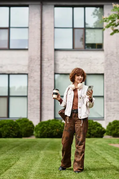 A young woman with curly hair stands outside her office, coffee in hand, checking her phone. — Stock Photo