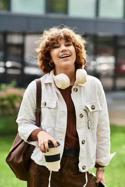 A cheerful young woman with curly hair stands near her office, sipping coffee and smiling. — Stock Photo