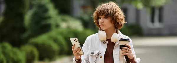 A young woman with curly hair holds a coffee cup and checks her phone near her modern workplace, banner — Stock Photo