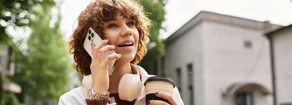 Curly-haired young woman talks on the phone, smiling and holding a coffee to go, banner — Stock Photo