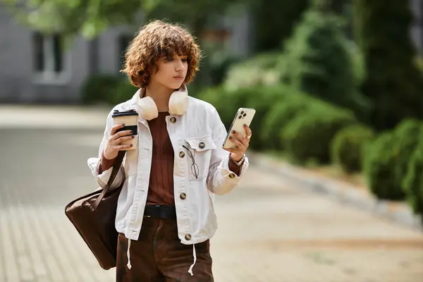 A young woman with curly hair walks near her office building, enjoying coffee and using her phone. — Stock Photo