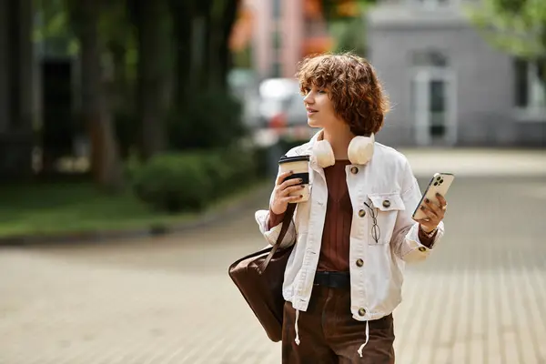 Curly-haired young woman relaxes outside her modern office, sipping coffee and checking her phone. — Stock Photo