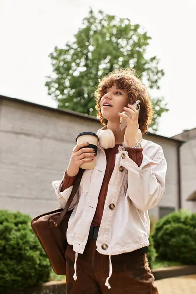 A young woman with curly hair chats on her phone, holding a coffee cup, near her office. — Stock Photo