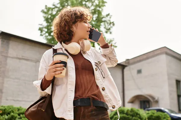 Curly-haired woman in casual attire holds coffee and phone near a sleek office structure. — Stock Photo