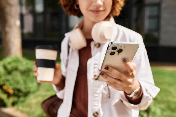 A young woman with curly hair holds a smartphone and coffee cup outside her office. — Stock Photo