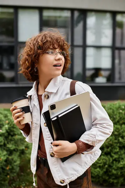 A young woman with curly hair holds a coffee and a laptop while standing near her workplace. — Stock Photo