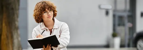 A young woman with curly hair sits writing in a notebook near a contemporary office building, banner — Stock Photo
