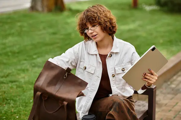 Curly-haired woman works remotely in a sunny park, balancing laptop and coffee. — Stock Photo
