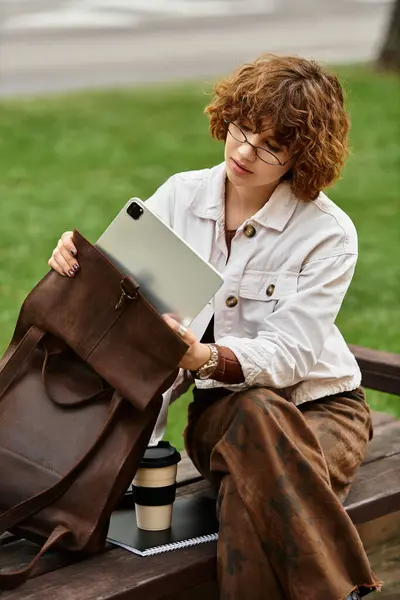 Young woman with curly hair works remotely on a tablet while seated on a blanket in summer sunshine. — Stock Photo