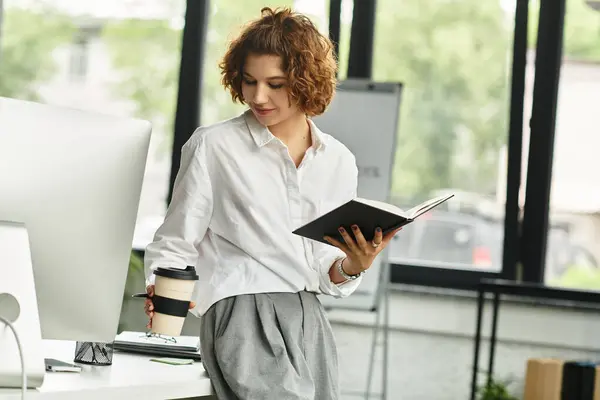 Curly-haired businesswoman reviews a notebook while holding a coffee cup in a bright office. — Stock Photo