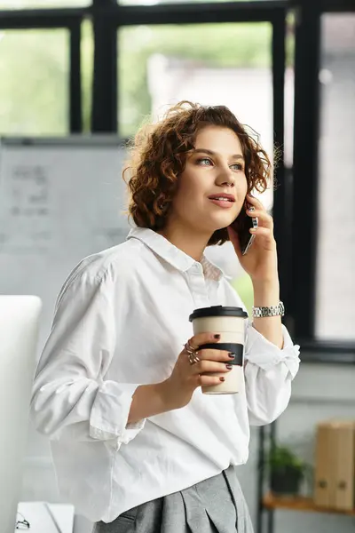 Young businesswoman with curly hair talks on the phone while holding coffee in a modern office. — Stock Photo