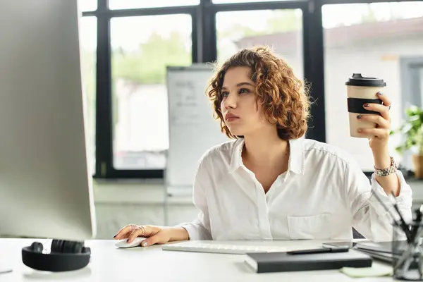 Curly-haired young woman engages with her computer while holding a cup of coffee in a sleek office. — Stock Photo