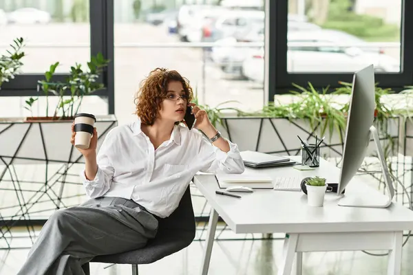 A young businesswoman with curly hair talks on the phone and enjoys her coffee at a desk. — Stock Photo