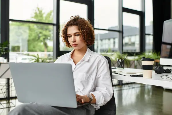 Curly-haired young businesswoman engages with her laptop in a contemporary office space. — Stock Photo