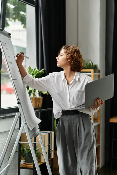 Businesswoman with curly hair actively writing on a whiteboard while holding a laptop. — Stock Photo