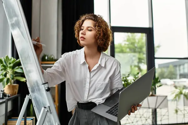 A young woman with curly hair actively works at a whiteboard while holding her laptop. — Stock Photo