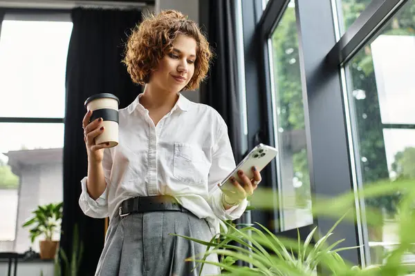 Curly-haired professional enjoys coffee while checking her smartphone in a bright office space. — Stock Photo