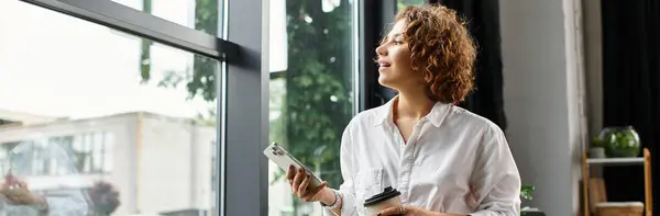 A young woman with curly hair stands by the office window, holding a phone and drink, banner — Stock Photo
