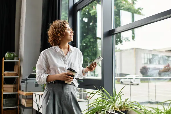 Young businesswoman with curly hair holds a coffee cup and looks out the window in her office. — Stock Photo